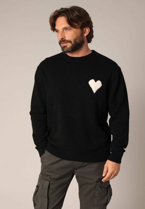Pull noir en matériau doux, avec un motif de cœur texturé blanc sur le côté gauche de la poitrine. Encolure et poignets côtelés.