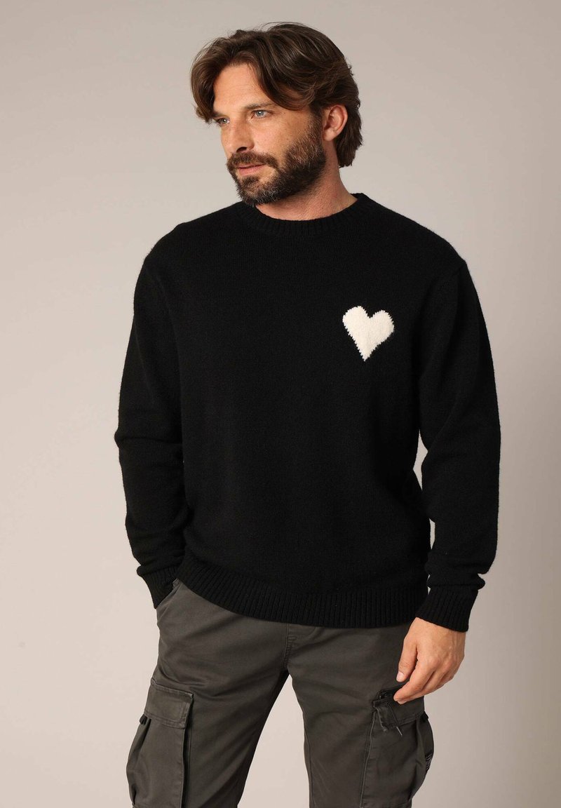 Zwarte trui van zacht materiaal, met een geborduurd wit hartmotief op de linkerborst. Ribbelkraag en -manchetten.