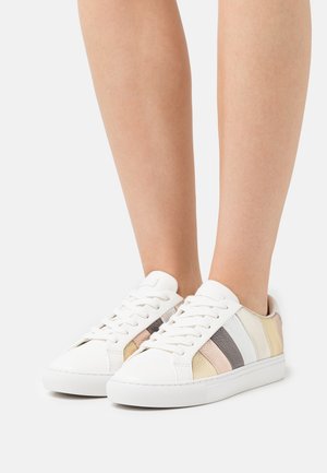 Hvide snøresneakers med flerfarvede pastelfarvede striber på siderne båret på bare ben, vist fra front- og sidevinkler.