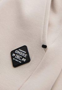 Patch noir en forme de diamant avec texte blanc « OMBRE INC. 85 » sur tissu beige avec couture visible et petit bouton noir.