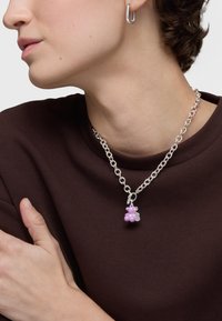 Srebrny naszyjnik łańcuszkowy z różowym uroczym misiem, okrągłymi kształtami i gładką teksturą, noszony na ciemno brązowej koszuli.