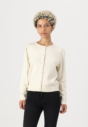 Cream-colored knit cardigan with a textured pattern, round neckline, button closure, and ribbed cuffs and hem. Worn with black jeans.