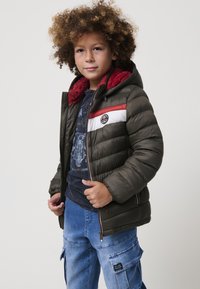 Niño con cabello rizado que lleva una chaqueta acolchada verde con un forro rojo, una camiseta gráfica azul marino y jeans cargo azules, de pie frente a un fondo liso.