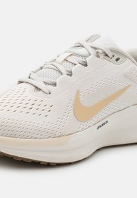 Pantof de sport bej deschis, cu un material superior din plasă respirabilă, detaliu auriu în formă de swoosh, talpă texturată și talpă intermediară cu amortizare, având inscripția "AIR".