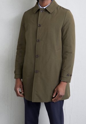 Homme portant un manteau vert olive boutonné avec des poches à rabat et un pantalon sombre, debout contre un mur uni.