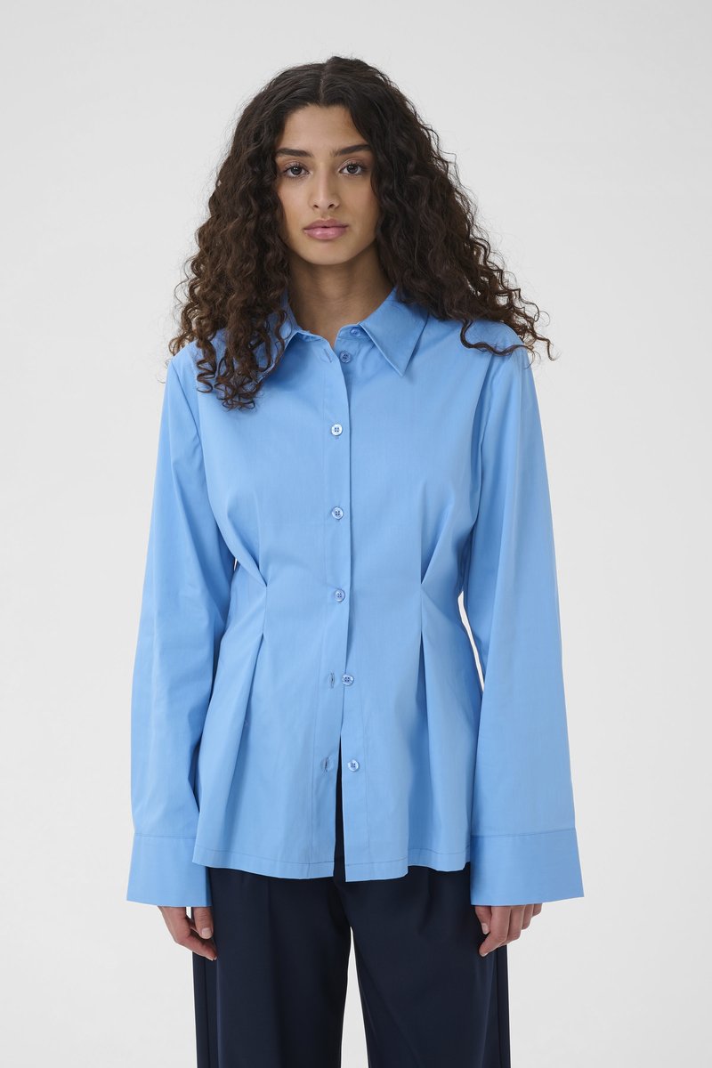 Kobieta o długich kręconych włosach, ubrana w jasnoniebieską koszulę zapinaną na guziki z szerokimi mankietami oraz czarne spodnie, stojąca na jednolitym tle.
