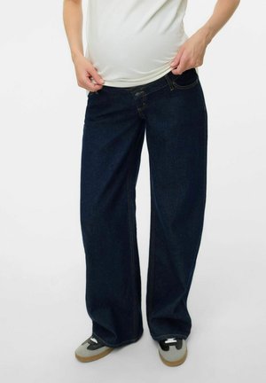 Jeans de pierna ancha de color azul oscuro en denim con costuras amarillas, cierre de botón y bolsillos frontales. Combinados con zapatillas grises.