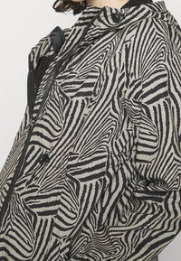 Primo piano di una persona che indossa una giacca a strisce zebra in bianco e nero con zip e cappuccio, mostrando dettagli di texture e pattern.