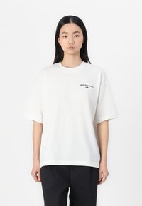 White cotton t-shirt with short sleeves, round neck, and a small black "New Balance" logo printed on the left side. Plain design.