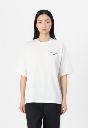 White cotton t-shirt with short sleeves, round neck, and a small black "New Balance" logo printed on the left side. Plain design.