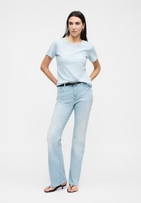 Frau steht mit den Händen hinter dem Rücken, trägt ein hellblaues T-Shirt, helle Jeans, schwarzen Gürtel, Creolen und schwarze Sandalen mit Absatz.