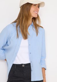 Light blue button-up shirt with long sleeves, made of smooth fabric, worn over a white t-shirt, paired with black pants.