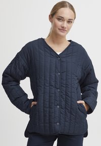 Mørkeblå quiltet jakke med v-hals, rund kant, frontlukning med knapper og sidelommer. Materialet ser blødt ud med en struktureret, puffet finish.