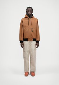 Καφέ τζάκετ με κουκούλα και μπροστινή τσέπη, μαύρες λεπτομέρειες, μπεζ cargo παντελόνια και πορτοκαλί παπούτσια με υφή.