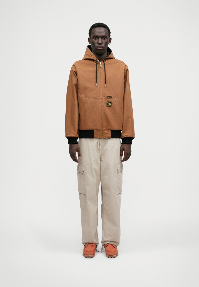 Καφέ τζάκετ με κουκούλα και μπροστινή τσέπη, μαύρες λεπτομέρειες, μπεζ cargo παντελόνια και πορτοκαλί παπούτσια με υφή.