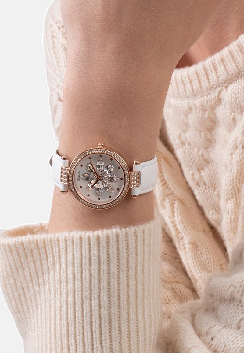 Valge rihmaga käekell roosa kuldse raamiga, millel on dialil kristalllillede disain, kantud vasakul käsivarrel kreemikuidas kampsuni peal.