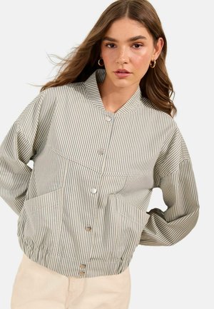 Junge Frau mit langen braunen Haaren trägt goldene Creolen und eine beige, vertikal gestreifte Jacke mit Metallknöpfen, posiert vor weißem Hintergrund.