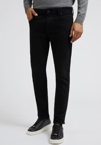 Fekete farmernadrág, slim fit fazonban, öt zsebes dizájnnal és finom textúrával. Fekete sneaker cipővel párosítva, fehér talppal.