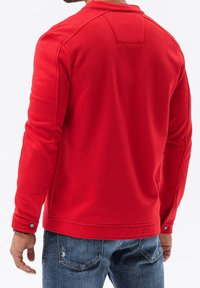 Felpa rossa con un tessuto morbido e testurizzato; presenta un colletto e dettagli con bottoni sui polsini. Indossata con jeans blu usurati.