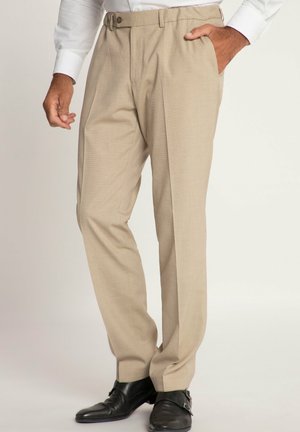 Mann trägt beige Stoffhose und schwarze Monk-Strap-Schuhe, Hand in der Tasche, steht vor einfachem Hintergrund.