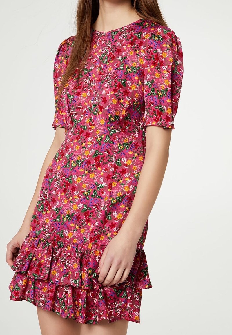 Mujer con vestido corto de manga corta, estampado floral rosa con borde con volantes, de pie frente a un fondo blanco liso.