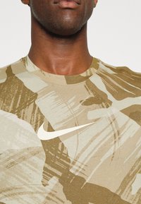 Camiseta de manga corta tan con patrones de pinceladas abstractas en verde y beige, con un logotipo de Nike en blanco en el pecho.