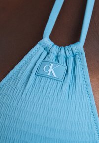 Kék nyakpántos bikini felső strukturált anyagból. Ráncolt anyag, vékony pántok és egy téglalap alakú logópatent található a közepén.
