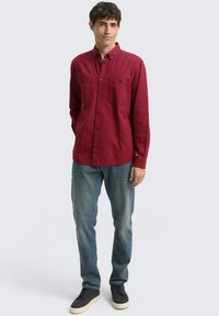 Chemise rouge à manches longues avec patte de boutonnage et deux poches poitrine, associée à un jean bleu clair et des chaussures foncées sur un fond neutre.