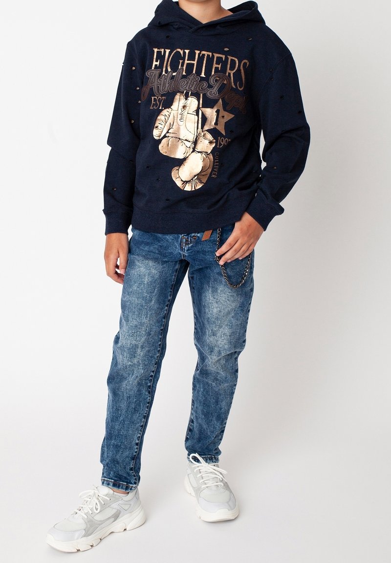 Sudadera de marina con diseño gráfico dorado de guantes de boxeo; combinada con jeans claros rasgados y zapatillas blancas con detalles en gris.