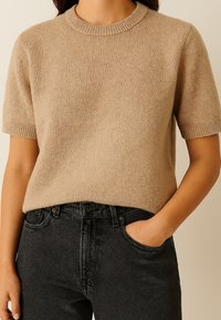 Beige trui met korte mouwen van gebreid materiaal, voorzien van een ronde hals en ribbelboorden. G gedragen bij hooggesneden donkere jeans. Zachte textuur.