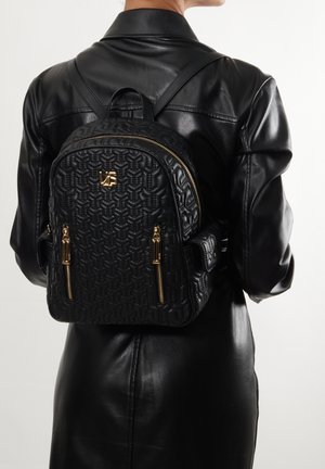 Mochila acolchada negra con herrajes dorados, que presenta dos cremalleras frontales y un asa superior. Llevada en la espalda, combinada con un conjunto de cuero negro.