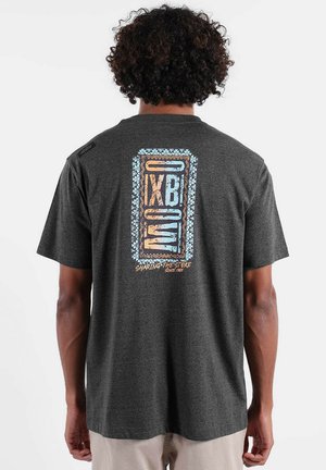 Personne aux cheveux bouclés portant un t-shirt gris foncé avec un logo géométrique bleu et orange et le texte "IXBOW Sharing the Stoke Since 1965" au dos.