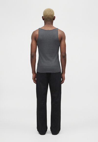 Άτομο με κοντά ξανθά μαλλιά στέκεται με την πλάτη γυρισμένη, φοράει αμάνικο γκρι πουκάμισο και φαρδιά μαύρα παντελόνια σε απλό φόντο.