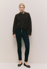 Ruskea, pörröinen neuletakki, jossa on napitus edessä ja kaksi taskua, yhdistettynä tummiin slim fit -farkkuihin ja mustiin leopardikuvioisiin ballerinoihin. Neutraali tausta.