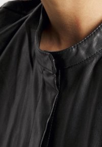 Svart läderplagg med hög krage, slät yta och en minimalistisk design med dold knappslå och subtila sömmar.