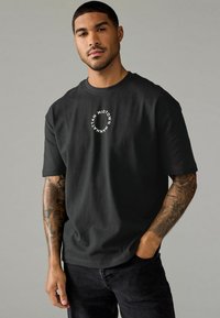 Sort bomulds T-shirt med et cirkulært hvidt tryk med teksten "Midtown Manhattan". Korte ærmer og afslappet pasform, parret med mørke jeans.