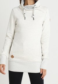 Vaaleanharmaa fleece-pullover, jossa on korkea kaulus, kuvioitu vuorikangas, nyörit ja pieni nahkainen logotarra edessä.