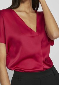 Blusa rossa in satin con scollatura a V, maniche corte e vestibilità rilassata. Caratterizzata da una texture liscia e un drappeggio sottile ai lati. Abbinata a pantaloni neri.