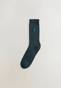 Mørkegrønne ribbede sokker med en glat tekstur og et lille blåt logo nær toppen. Forlænget, standard sokkeform.