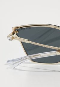 Kullatud prillid, millel on kuldne metallraam, mustad läätsed, selged templid ja kristallaktsent käepidemel, millel on "Swarovski" logo.