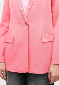 Blazer rose avec un bouton et deux poches avant à rabat porté sur un haut blanc et un pantalon gris foncé, mains reposant sur les côtés.