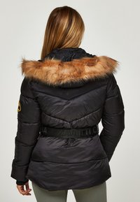 Chaqueta acolchada negra con capucha con borde de piel sintética, cintura ceñida y diseño acolchado. Presenta un parche con el logo en la manga izquierda.