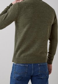 Pull tricoté vert olive avec une finition texturée, encolure ronde et poignets côtelés, associé à un jean en denim bleu avec poches arrière.