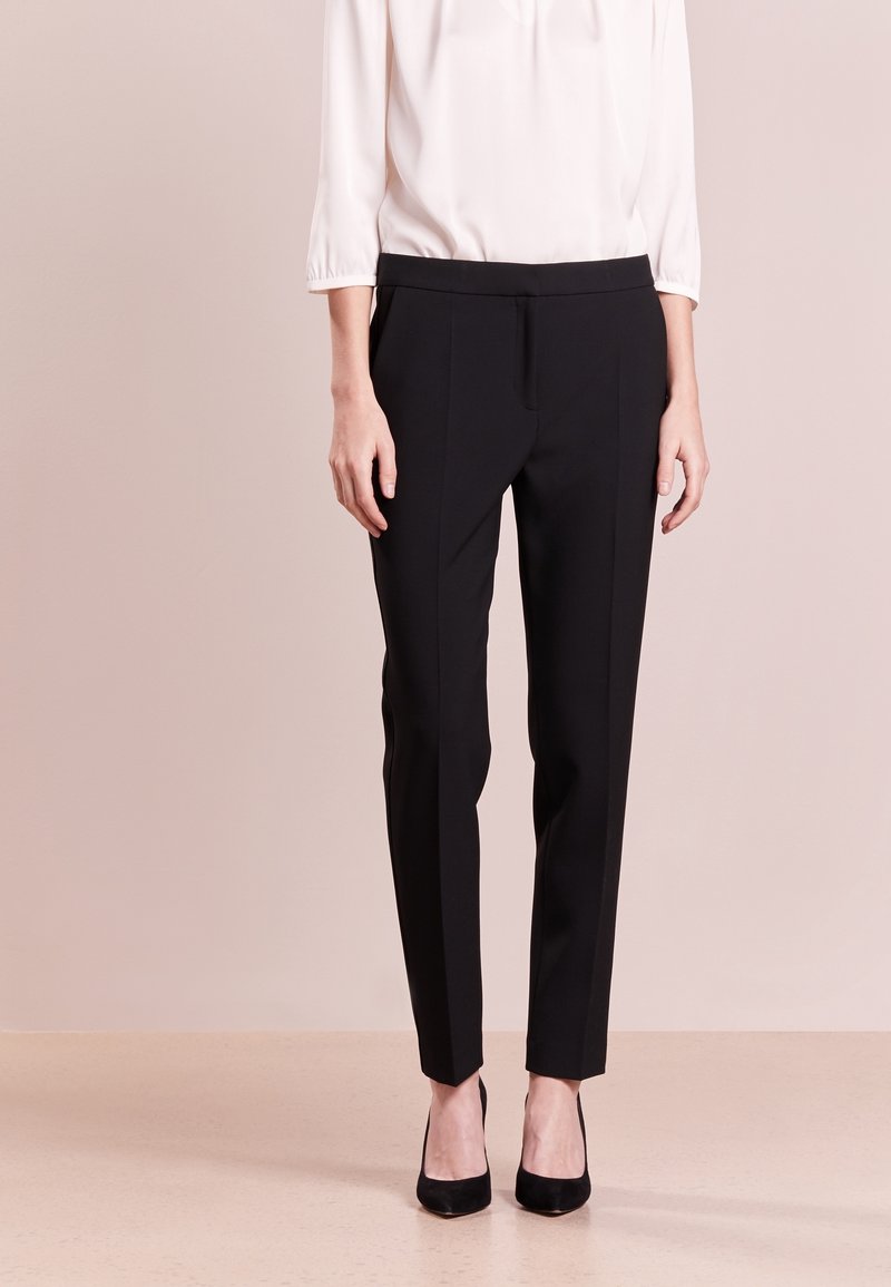 Femme portant un pantalon noir sur mesure et des talons noirs, associé à un chemisier blanc à manches longues, debout devant un fond uni.