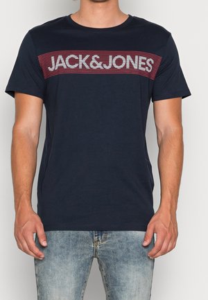 Mężczyzna w granatowym t-shircie z napisem "JACK & JONES" białymi literami na czerwono-paskowanym pasku, w zestawie z jasnoniebieskimi dżinsami.