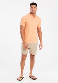 Heren polo met korte mouwen in koraal oranje, gemaakt van katoen. Gemaakt met beige shorts en bruine slippers. Eenvoudig ontwerp van de kraag.