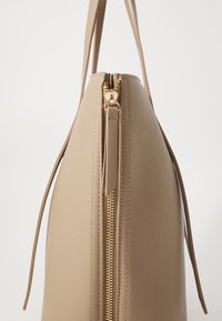 Beige håndtaske med en glat tekstur, en guld lynlås og et minimalistisk design. Har smalle stropper og en tilspidset silhuet.
