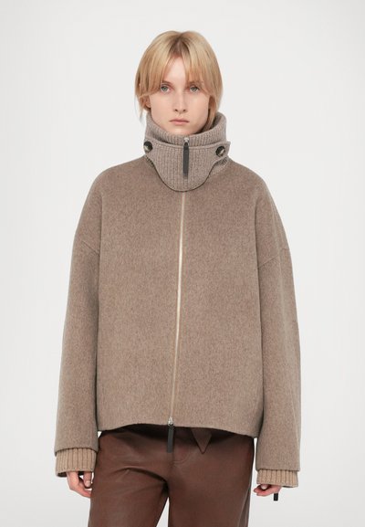 Übergroße beige Wolljacke mit hohem Kragen, ausgestattet mit einem Frontreißverschluss, gerippten Bündchen und schwarzen Hardware-Details. Glatte Textur.