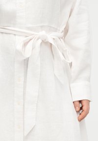 Λευκό φόρεμα πουκάμισο με μακριά μανίκια, κουμπιά μπροστά και δεμένη υφασμάτινη ζώνη στη μέση, με εμφανές το δεξί χέρι και την παλάμη.