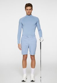 Vaaleansininen pitkähihainen paita ja siihen sopivat shortsit, molemmat sileää kangasta; golfaaja, joka pitää mailaa, pukeutuneena valkoisiin lenkkareihin ja käsineisiin.